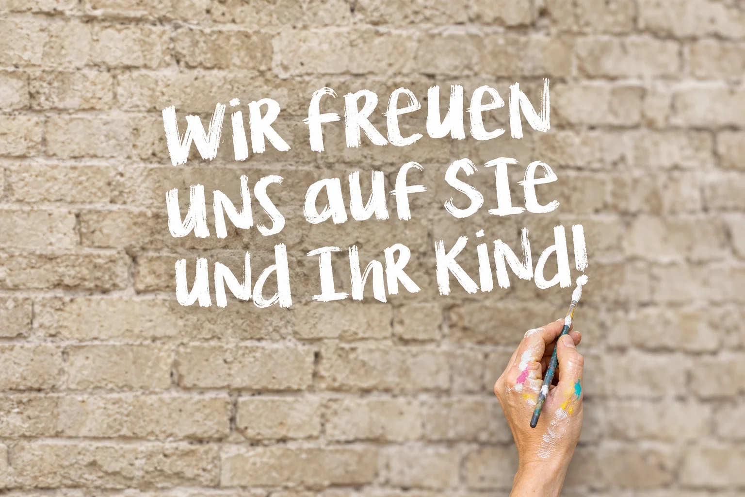 Wir freuen uns auf Sie und Ihr Kind! – weiße Pinselbuchstaben, die eine Hand mit einem Pinsel auf die Backsteinwand malt